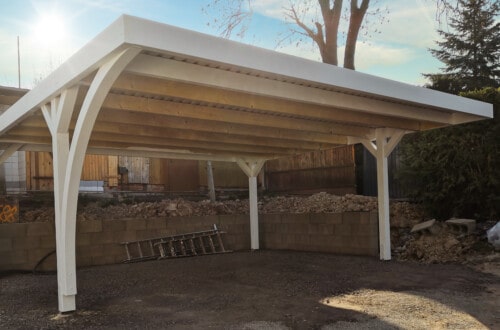 Carport A white, wooden carport with a slanted roof stands on a gravel surface, supported by four posts. There is a cinderblock wall behind it, a ladder, dirt piles, trees, and a house in the background. www.commaik.de