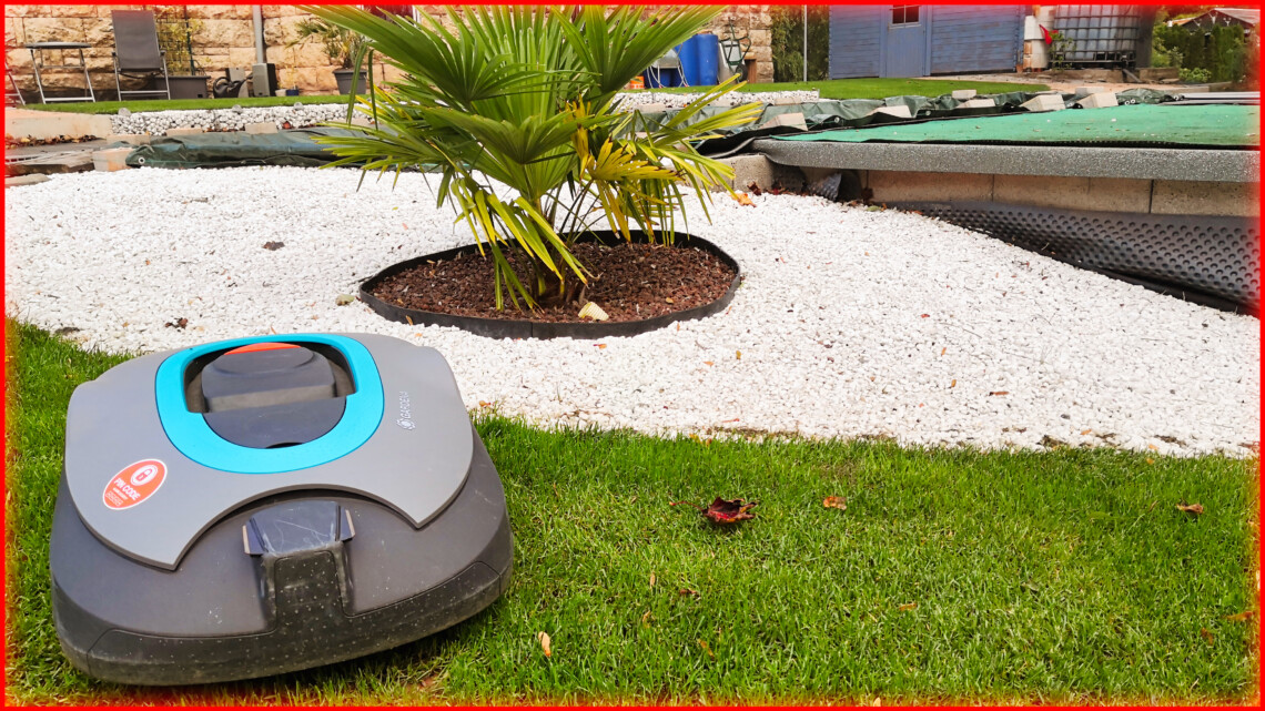 Garten 10 Ein Rasenroboter sitzt auf grünem Gras in der Nähe einer kleinen Palme, umgeben von weißem Kies in einem angelegten Garten, bereit zum Auswintern für die neue Mähsaison. www.commaik.de