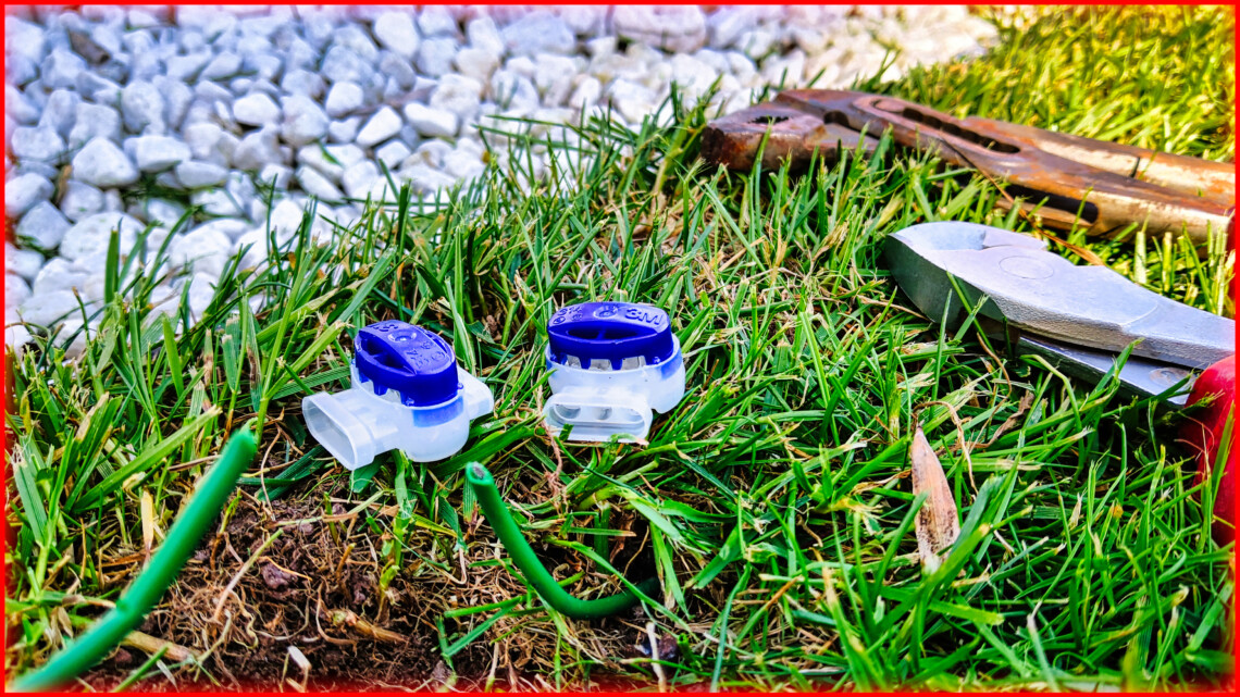 Garten 9 Zwei blaue und durchsichtige Kabelverbinder und grüne Drähte auf Rasen, mit Zange und weißem Kies im Hintergrund-unverzichtbare Werkzeuge, um Kabelbruch für Ihren Rasenroboter effizient zu reparieren. www.commaik.de