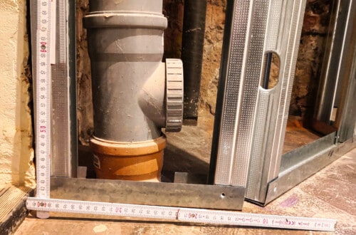 Tutorials A metal frame and a measuring tape are positioned around a large PVC pipe with a connector at its base, set against a concrete wall in a construction area. www.commaik.de