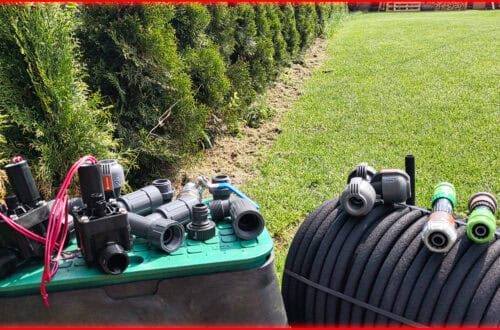 Verschiedene Teile des Bewässerungssystems und Anschlüsse für die Heckenbewässerung sind auf einem grünen Kasten neben einem aufgerollten schwarzen Gartenschlauch platziert, mit einem ordentlich getrimmten Rasen und einer Reihe von immergrünen Sträuchern im Hintergrund. www.commaik.de
