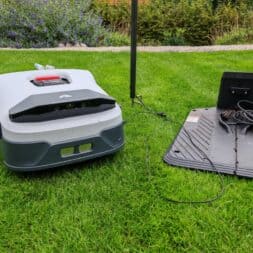 Anthbot Genie robotic lawnmower in test: Perfect lawn mowing without cables? 19 A robotic lawnmower sits on green grass near its charging station, focusing on how it works. A garden with shrubs and flowers can be seen in the background, emphasizing the intended use in a beautifully manicured garden. www.commaik.de