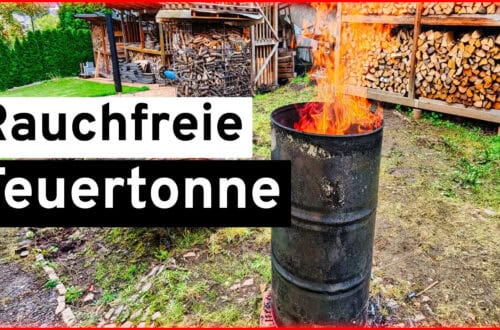 DIY: Build your own smoke-free fire barrel from an oil barrel A large oil barrel serves as a smoke-free fire barrel, burning with tall flames in a backyard with stacked firewood and sheds in the background. German text reads Rauchfreie Feuertonne. www.commaik.de