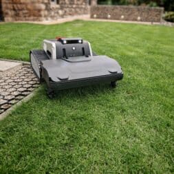 Lymow One in test: Robot mower without boundary wire with RTK navigation and chain drive 7 A Lymow One robotic lawnmower with tracks mows grass on a manicured lawn near a stone path and a building wall in the background. www.commaik.de