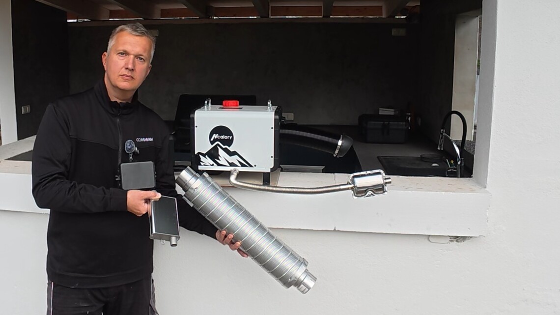 Eine Person in schwarzer Jacke steht vor einem weißen Tresen, in der Hand ein großes Metallauspuffrohr und ein Steuergerät mit Display. Auf dem Tresen steht eine Schachtel mit der Aufschrift Autoterm Standheizung und verschiedenen mechanischen Heizungsteilen. www.commaik.de