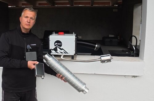 Heating optimization: How to make your diesel parking heater quieter - pump and silencer in the test 2 A person in dark clothing stands by a white counter, holding a metal tube and a small rectangular device. Behind the counter is a white and black parking heater machine labeled Motour with various attachments and a red button. www.commaik.de
