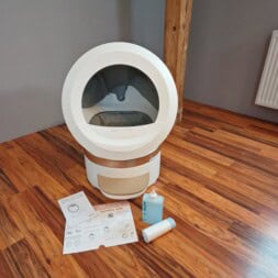 Automatic litter box test: Self-cleaning litter box without odor - Mova LR10 6 A white automatic self-cleaning litter box sits on a wooden floor. In front of the litter box are instruction sheets, a bottle of cleaner, and a roll of waste bags. The background features a radiator and a watering can. www.commaik.de