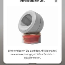 Automatic litter box test: Self-cleaning litter box without odor - Mova LR10 78 A smartphone screen displays a litter box app notification: "Abfallbehälter voll" (waste bin full), with an image of a round automatic litter box and instructions to empty the bin for proper operation. www.commaik.de