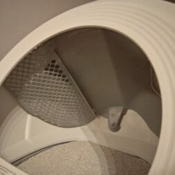 Automatic litter box test: Self-cleaning litter box without odor - Mova LR10 45 Close-up view of the interior of a washing machine, showing the drum and a section of the lint filter with perforated metal—resembling the texture found in some litter box designs. Shadows and curved white plastic edges are also visible. www.commaik.de