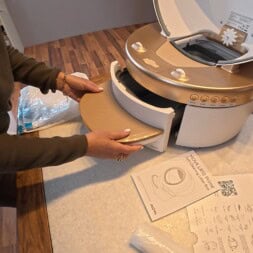 Automatic litter box test: Self-cleaning litter box without odor - Mova LR10 26 A person assembles a gold and white litter box on a countertop, sliding its drawer into place. Instruction manuals, plastic components, and packaging materials are visible nearby. www.commaik.de