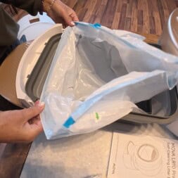 Automatic litter box test: Self-cleaning litter box without odor - Mova LR10 28 A person places a plastic liner inside a rectangular litter box, which sits on a counter next to a white appliance and an instruction sheet with text and diagrams. Wood flooring is visible in the background. www.commaik.de