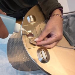 Automatic litter box test: Self-cleaning litter box without odor - Mova LR10 31 A person fills a small container on a gold-colored toilet with blue liquid from a squeeze bottle, possibly for cleaning or maintenance. The toilet lid is open, and a trash bag—much like in a litter box—is visible inside. www.commaik.de