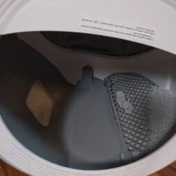 Automatic litter box test: Self-cleaning litter box without odor - Mova LR10 35 Open front-loading dryer with a gray lint trap partially removed, containing some lint and debris—similar to cleaning a litter box. The dryer sits on a wooden floor, door open and the interior visible. www.commaik.de