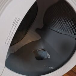 Automatic litter box test: Self-cleaning litter box without odor - Mova LR10 37 Close-up of the open door of a front-loading clothes dryer, showing the empty drum and lint filter inside. A gray mat is nearby on the wooden floor, next to a litter box placed discreetly out of direct view. www.commaik.de
