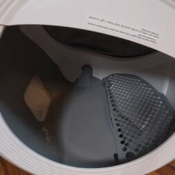 Automatic litter box test: Self-cleaning litter box without odor - Mova LR10 38 The image shows the open door of a clothes dryer, displaying the drum and a removable lint filter, with the appliance sitting on a wooden surface. A litter box is visible nearby, but the dryer itself appears empty. www.commaik.de