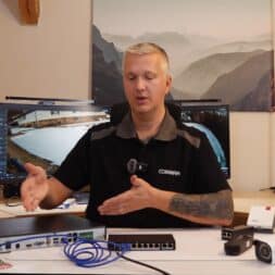 Install surveillance cameras yourself - IP camera network explained simply 37 A man is sitting at a desk with various surveillance and network devices, including a surveillance camera, an IP camera, a router and cables. He demonstrates their use, while two monitors behind him display the footage from the surveillance camera. www.commaik.de