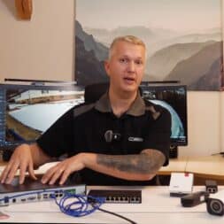 Install surveillance cameras yourself - IP camera network explained simply 46 A man sits at a desk with various surveillance cameras and network devices. Behind him, two monitors show a surveillance image and a mountain landscape. It seems to explain the devices. www.commaik.de