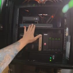 Install surveillance cameras yourself - IP camera network explained simply 40 A tattooed arm reaches into a server rack filled with network devices - including routers, NAS devices and an IP camera - and illuminated by LED lights. www.commaik.de