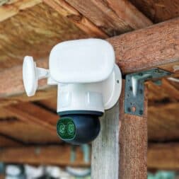 The best Reolink 4k PTZ surveillance cameras in comparison: 823S2 vs 823S1 vs TrackMix vs TrackFlex in test 28 A white Reolink security camera with two lenses, 4K PTZ capability and a floodlight is mounted on a wooden beam under a roof, facing outwards for surveillance. www.commaik.de
