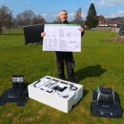 Lymow One Plus in test: RTK robot mower for large areas & difficult terrain 9 A person stands in a meadow holding a large instruction sheet surrounded by parts of the Lymow One Plus robotic lawnmower, including base plates, accessories in a foam tray and the mower itself, against a rural background. www.commaik.de