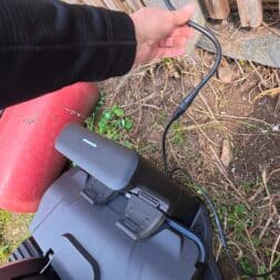 Lymow One Plus in test: RTK robot mower for large areas & difficult terrain 13 A person holds a charging cable in his hand and connects it to the battery of a Lymow One Plus electric lawnmower standing on the ground next to a wooden fence. A red plastic container and some plants can be seen nearby. www.commaik.de