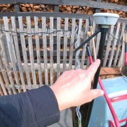 Lymow One Plus in test: RTK robot mower for large areas & difficult terrain 14 A hand points to a black metal pole with a white power box attached to it, possibly for a Lymow One Plus, in front of a wooden fence with firewood stacked on top. Various objects, including a red trolley and a blue garbage can, are standing nearby. www.commaik.de