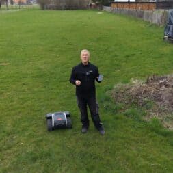 Lymow One Plus in test: RTK robot mower for large areas & difficult terrain 27 A man stands in a meadow holding a smartphone next to the Lymow One Plus robotic lawnmower. To his right is a pile of rubble and wooden planks, with fences and buildings in the background. www.commaik.de