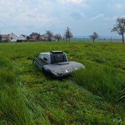 Lymow One Plus in test: RTK robot mower for large areas & difficult terrain 35 A Lymow One Plus robotic lawnmower moves through tall green grass in a large field, with houses, trees and a cloudy sky in the background. www.commaik.de