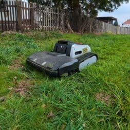 Lymow One Plus in test: RTK robot mower for large areas & difficult terrain 36 A Lymow One Plus robotic lawnmower with tank-like tracks sits on tall, green grass in a garden near a wooden fence, with houses and trees visible in the background. www.commaik.de