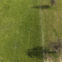 Lymow One Plus in test: RTK robot mower for large areas & difficult terrain 30 Aerial view of a grassy field divided by a faint fence line, with scattered trees casting shadows. In the center of the field is a Lymow One Plus, while a trampoline can be seen in the top left corner. www.commaik.de