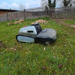 Lymow One Plus in test: RTK robot mower for large areas & difficult terrain 37 A Lymow One Plus robotic lawnmower drives across a grassy yard with scattered pieces of wood nearby, bordered by a wooden fence and trees in the background. www.commaik.de