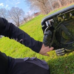Lymow One Plus in test: RTK robot mower for large areas & difficult terrain 41 A person in black clothing kneels in the grass and checks the wheel and caterpillar mechanism of a Lymow One Plus robotic lawnmower under a bright, partly cloudy sky. Trees and a fence can be seen in the background. www.commaik.de
