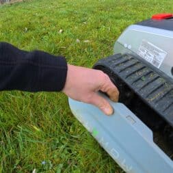 Lymow One Plus in test: RTK robot mower for large areas & difficult terrain 49 A person's hand lifts the cover of a Lymow One Plus robotic lawnmower on green grass, revealing the internal chains and components. www.commaik.de