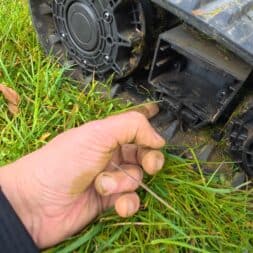 Lymow One Plus in test: RTK robot mower for large areas & difficult terrain 51 Close-up of a hand repairing or inspecting the underside of a Lymow One Plus outdoor appliance, possibly a lawn mower, on green grass. The hand holds a part near the wheel and gear parts of the machine. www.commaik.de