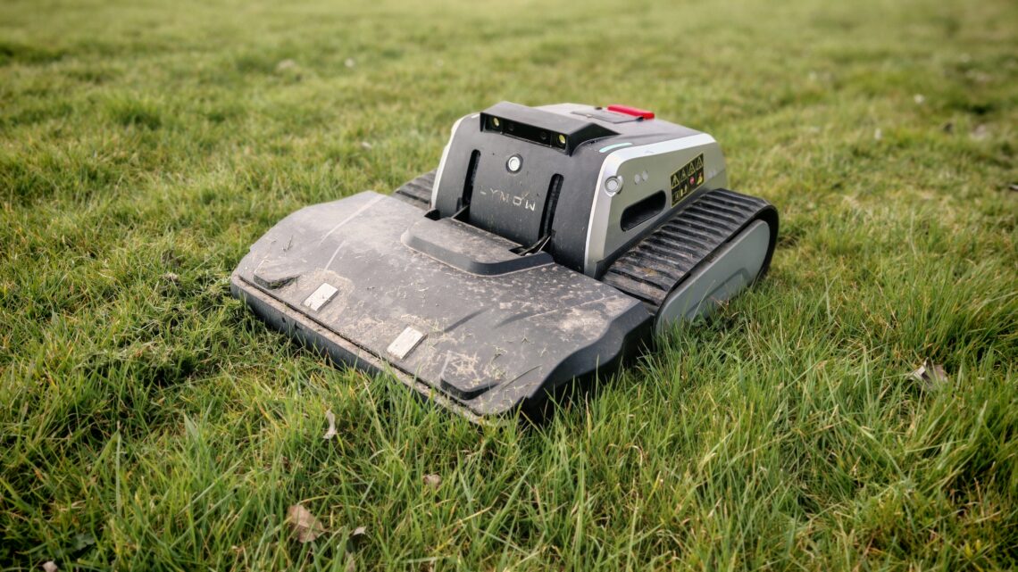 Ein Lymow One Plus Rasenmähroboter mit panzerähnlichen Raupen sitzt auf grünem Gras, leicht verschmutzt vom Gebrauch, in einem Außenbereich. www.commaik.de