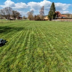 Lymow One Plus in test: RTK robot mower for large areas & difficult terrain 3 A large grass field with a Lymow One Plus robotic lawnmower in the foreground, under a blue sky with scattered clouds. Trees and houses can be seen in the background. www.commaik.de