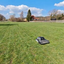 Lymow One Plus in test: RTK robot mower for large areas & difficult terrain 5 A Lymow One Plus robotic lawnmower drives across a large, green field of grass under a blue sky with scattered clouds. Trees and houses can be seen in the background. www.commaik.de