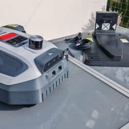 Robot lawn mower without boundary wire in test: LiDAR vs RTK in comparison (ANTHBOT M5 vs M9) 5 An Anthbot robotic lawnmower and its charging station stand on a glass surface outside, in front of a white wall and a wire fence in the background. www.commaik.de