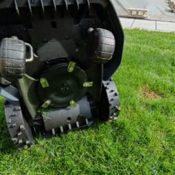 Robot lawn mower without boundary wire in test: LiDAR vs RTK in comparison (ANTHBOT M5 vs M9) 10 Close-up of the underside of an Anthbot robotic lawnmower on grass, highlighting the wheels, cutting blades and treads. A terrace and a lawn can be seen in the background. www.commaik.de