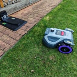 Robot lawn mower without boundary wire in test: LiDAR vs RTK in comparison (ANTHBOT M5 vs M9) 17 An Anthbot robotic lawnmower stands in the meadow near a paved area and drives to its charging station by a pile of wood along a wooden wall. www.commaik.de