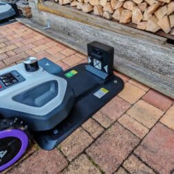 Robot lawn mower without boundary wire in test: LiDAR vs RTK in comparison (ANTHBOT M5 vs M9) 18 An Anthbot robotic mower with purple wheels docks at a charging station on a brick terrace next to a pile of wood; another similar mower can be seen in the background. www.commaik.de