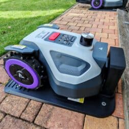 Robot lawn mower without boundary wire in test: LiDAR vs RTK in comparison (ANTHBOT M5 vs M9) 19 An Anthbot robotic mower with a gray and black body and purple wheels stands at a charging station near a meadow and a pile of firewood on a brick patio. www.commaik.de
