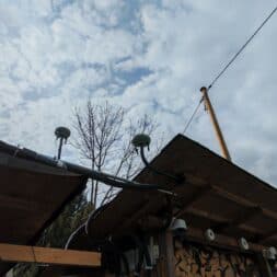 Robot lawn mower without boundary wire in test: LiDAR vs RTK in comparison (ANTHBOT M5 vs M9) 23 Low view of wooden roofs with satellite dishes, antennas and a robot mower charging station against a cloudy sky. Stacked firewood can be seen under the roof, with tall trees and a solar panel in the background. www.commaik.de