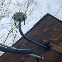 Robot lawn mower without boundary wire in test: LiDAR vs RTK in comparison (ANTHBOT M5 vs M9) 24 A close-up of a satellite antenna mounted on the underside of a wooden roof, with cables attached and bare tree branches in the background against a cloudy sky - an ideal location for an anthbot or robotic mower signal receiver. www.commaik.de