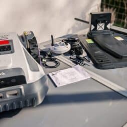 Robot lawn mower without boundary wire in test: LiDAR vs RTK in comparison (ANTHBOT M5 vs M9) 22 An Anthbot robotic lawnmower, its charging station, power cable, small accessories and a user manual lie on a glass table outside. www.commaik.de