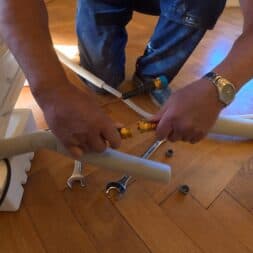 Installing Air Conditioning Yourself in an Older Building – Heating and Cooling in Practice 25 A person is installing an air conditioner and using wrenches to connect insulated pipes on a wooden floor. Tools and installation materials are scattered around nearby. www.commaik.de