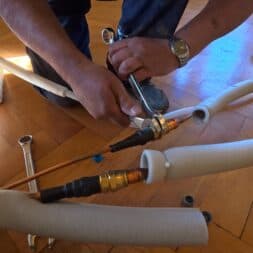 Installing Air Conditioning Yourself in an Older Building – Heating and Cooling in Practice 27 A person is using a wrench to tighten copper pipes connected to an air conditioner; foam insulation and tools are visible on the wooden floor. www.commaik.de