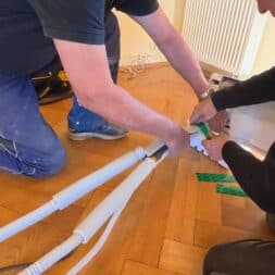 Installing Air Conditioning Yourself in an Older Building – Heating and Cooling in Practice 28 Two people are kneeling on a wooden floor, laying insulated HVAC pipes, possibly for an air conditioning or heating system. One person holds the wires in place while the other wraps them with tape; tools and components are scattered nearby. www.commaik.de