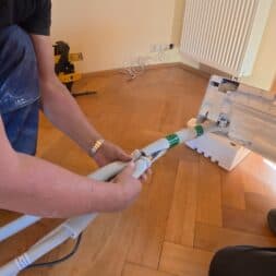 Installing Air Conditioning Yourself in an Older Building – Heating and Cooling in Practice 29 Two people are installing or connecting a white plastic hose to an indoor air conditioner mounted on the wall; tools and cables are visible on the wooden floor. www.commaik.de