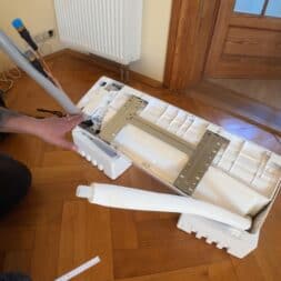 Installing Air Conditioning Yourself in an Older Building – Heating and Cooling in Practice 22 A person is assembling or unpacking a white air conditioner unit—with metal brackets and foam packaging—on a wooden floor next to a door and a radiator. Tools and cables can be seen in the background. www.commaik.de